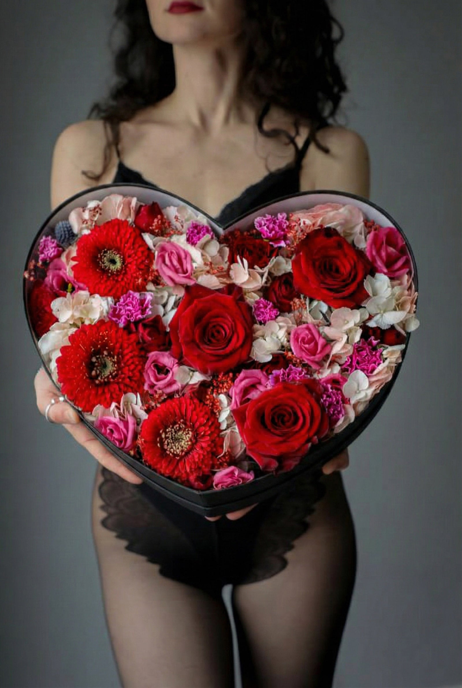 Heart-shaped box of red and pink flowers held by a person against a gray background
