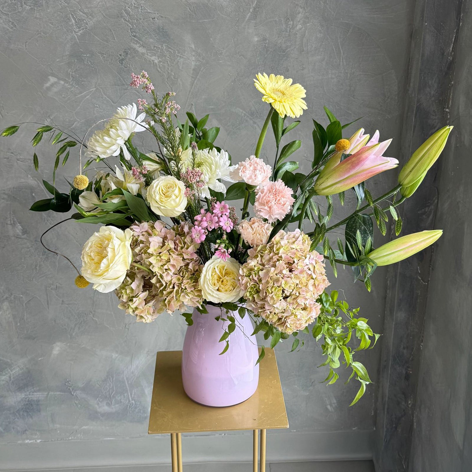 Bouquet of flowers in a pink vase on a gold stand against a gray wall.