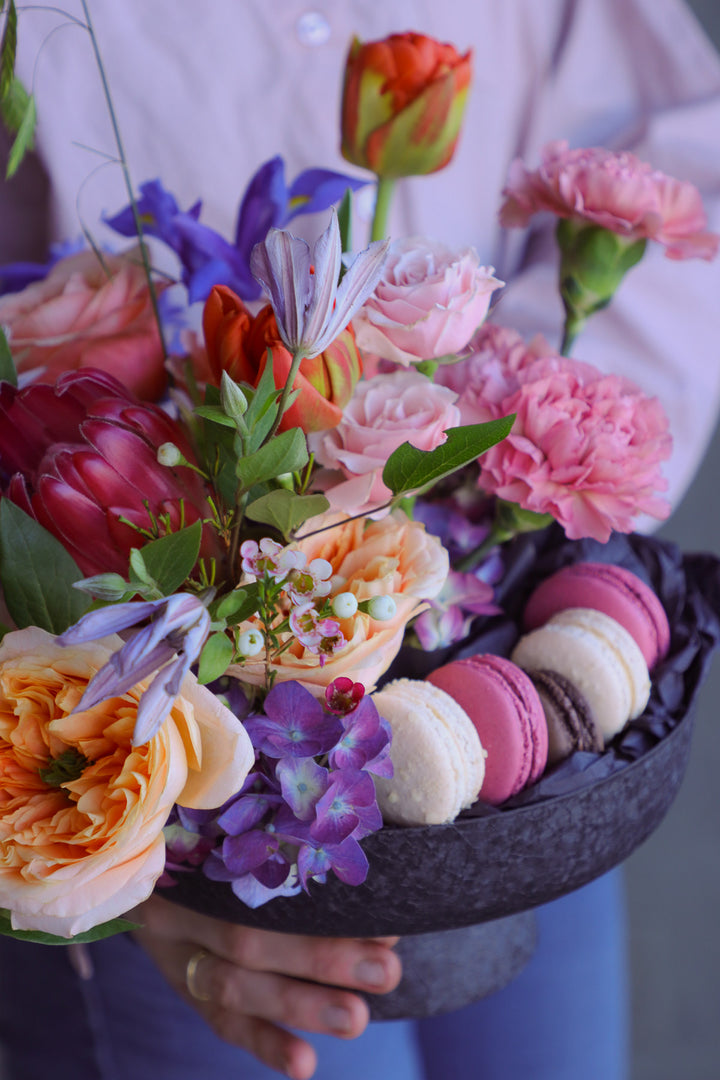 Colorful garden-style floral display in a dark pedestal bowl with pastel macarons.