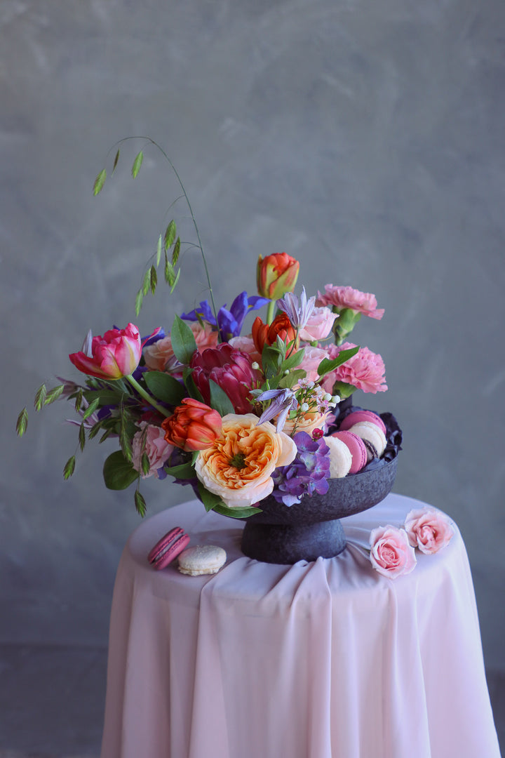 Bright summer flower arrangement with macarons in a black compote dish on draped fabric.