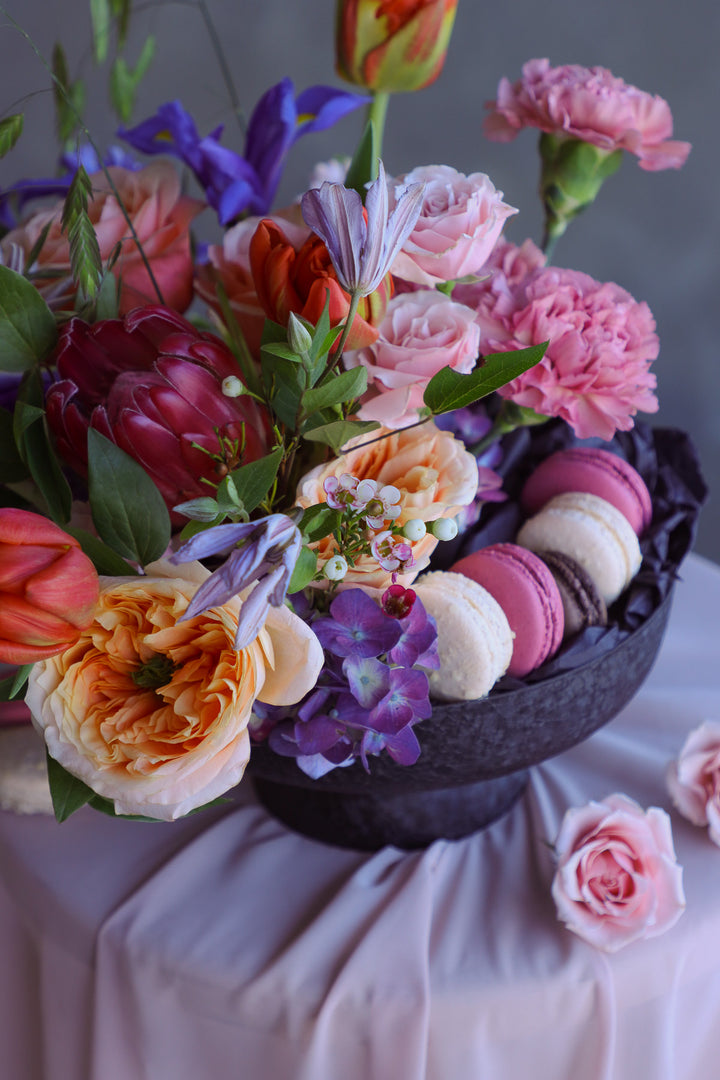 Bold pink, coral, and purple flower arrangement with macarons in a pedestal dish.