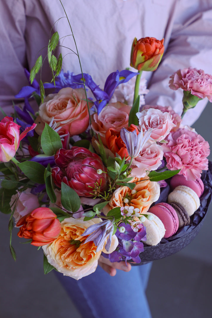 Close view of a person holding a colorful floral and macaron arrangement in a dark compote bowl.