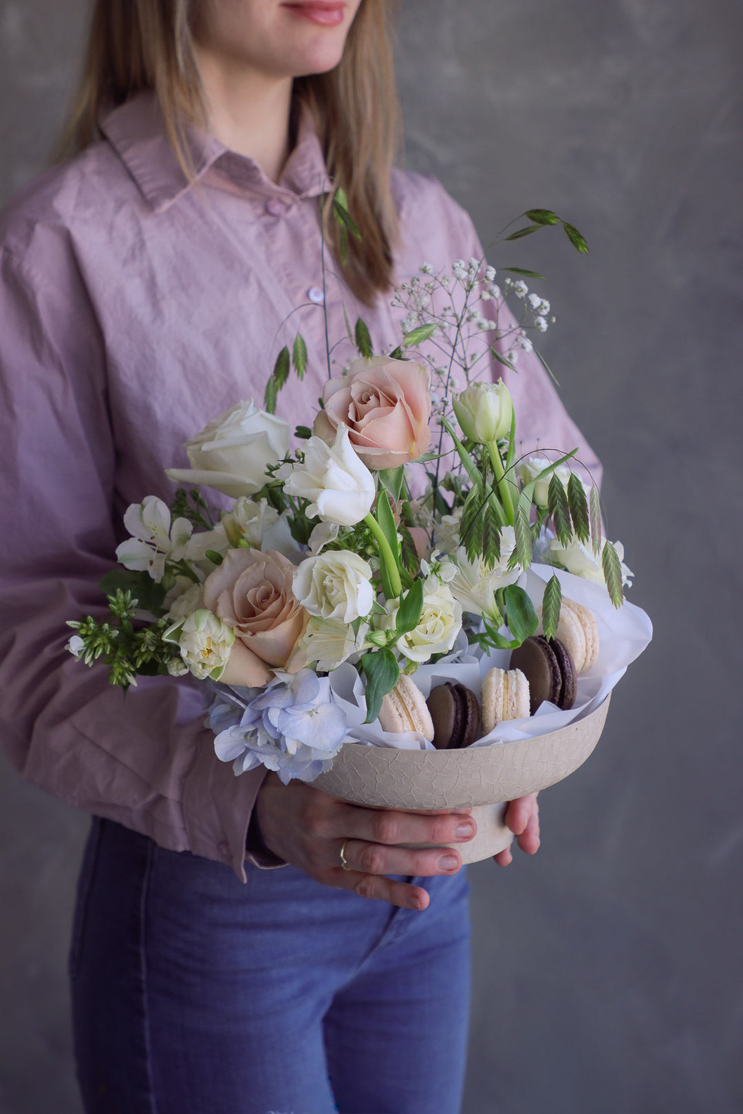Woman holding a garden-style arrangement in blush, cream, and pale blue tones with macarons nestled inside a pedestal bowl