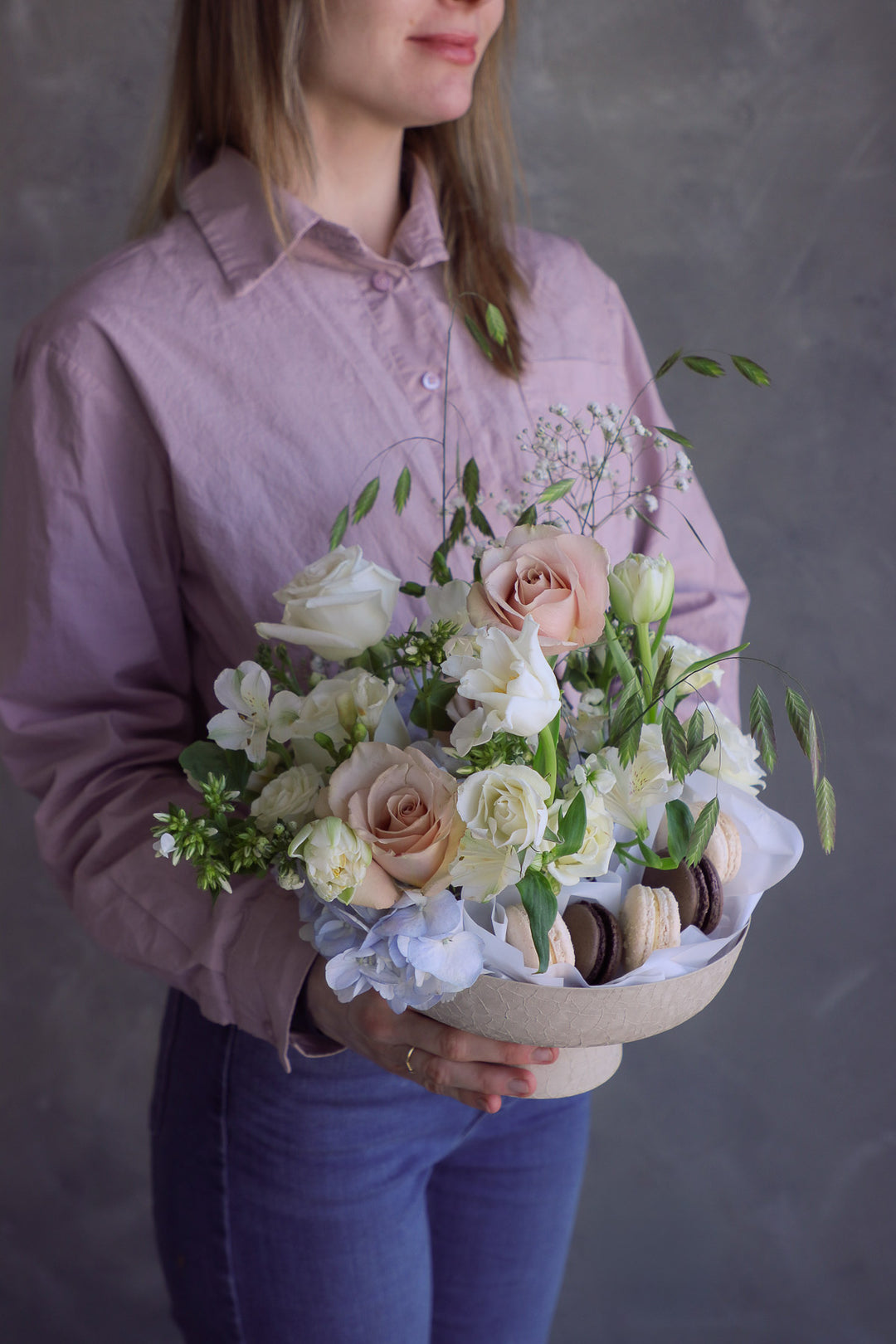 Woman holding a soft romantic floral arrangement with creamy blooms and macarons styled in a ceramic pedestal dish.