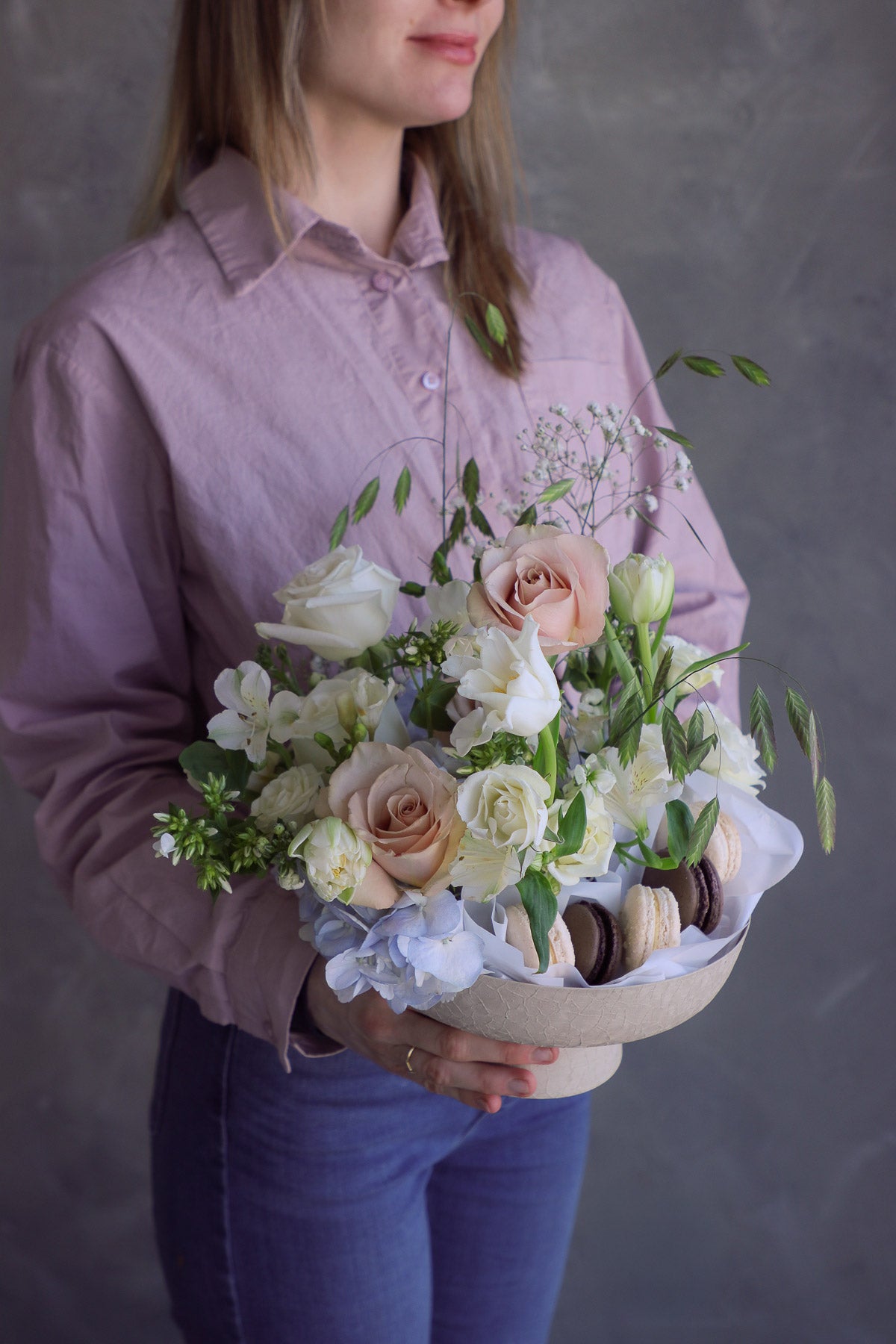 Woman holding a soft romantic floral arrangement with creamy blooms and macarons styled in a ceramic pedestal dish.