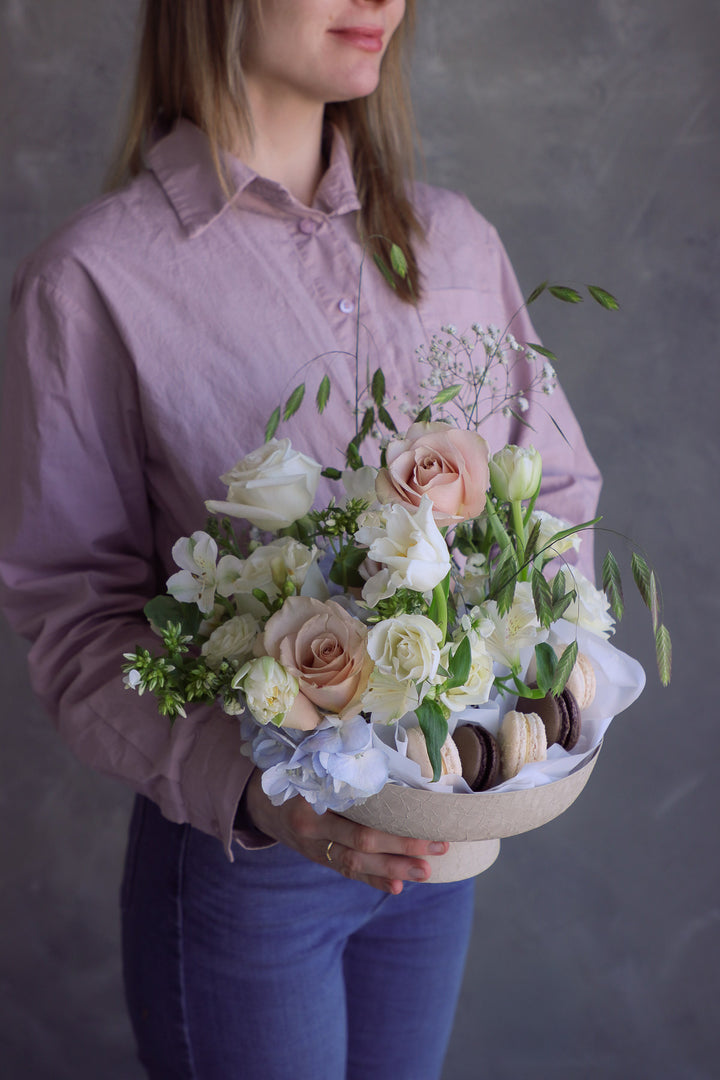 Woman holding a soft romantic floral arrangement with creamy blooms and macarons styled in a ceramic pedestal dish.
