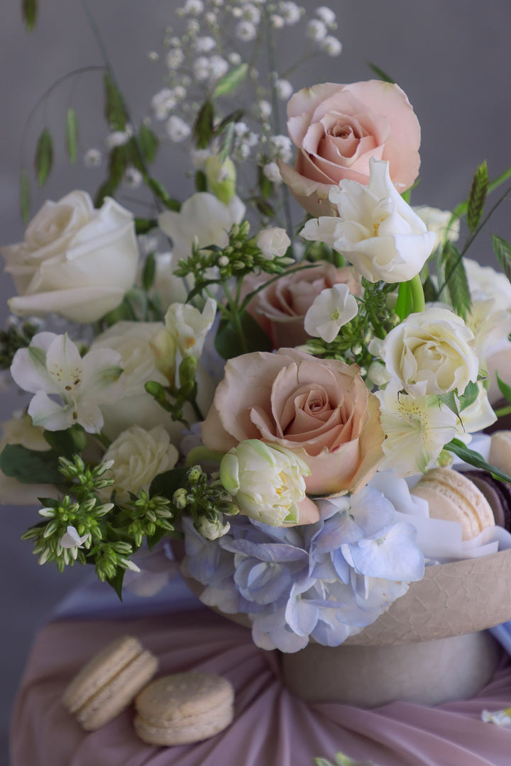 Creamy white and soft blue floral arrangement with macarons in a nude ceramic compote dish on draped fabric.