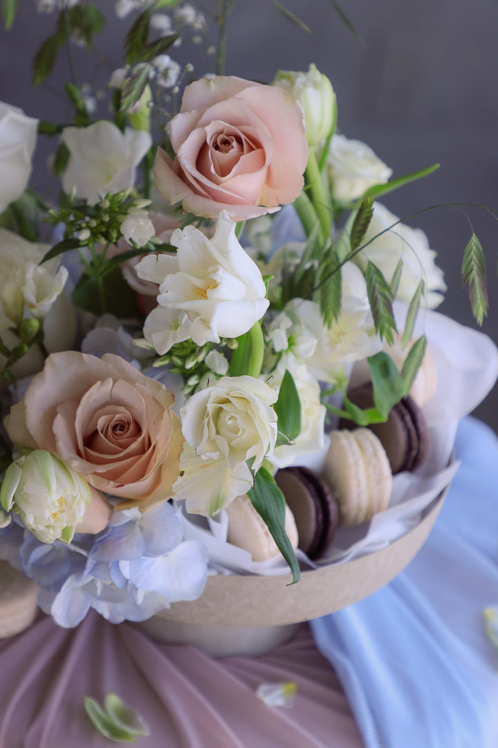 Garden-style arrangement in blush, cream, and pale blue tones with macarons nestled inside a pedestal bowl.