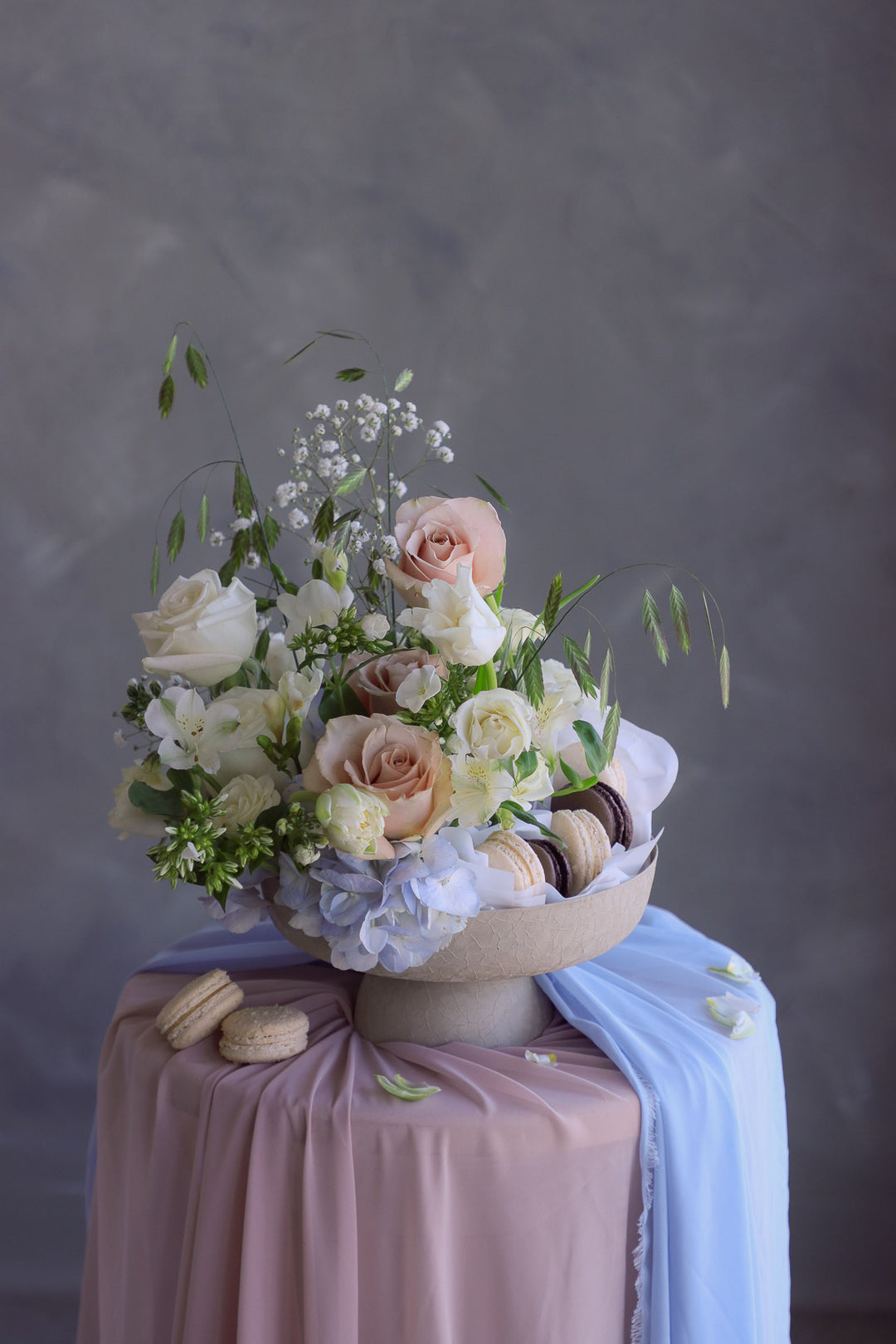 Elegant pastel flower display in a textured nude compote with delicate greenery and French macarons on a draped pedestal