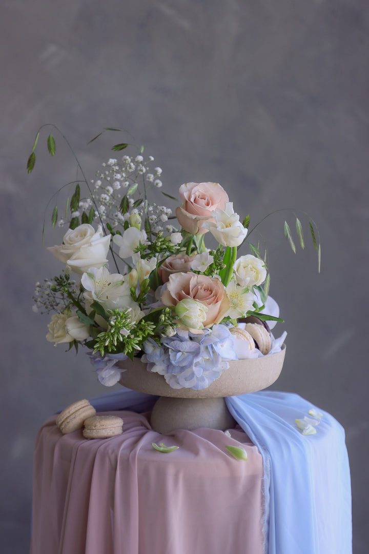 Soft romantic floral arrangement with creamy blooms and macarons styled in a ceramic pedestal dish.