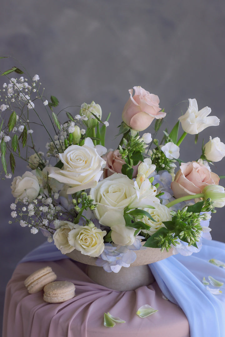 Cream-toned flowers with soft blue accents arranged in a raised ceramic dish with macarons tucked inside.