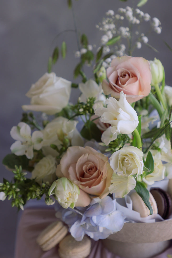 Close up of cream-toned flowers with soft blue accents arranged in a raised ceramic dish with macarons tucked inside.