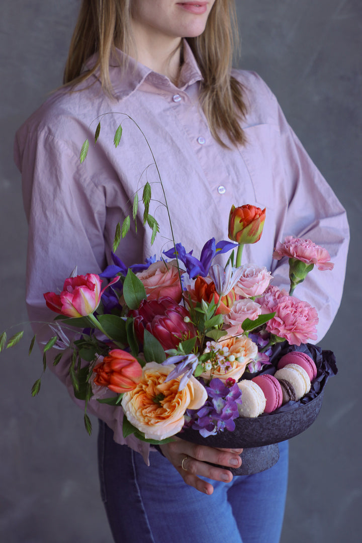 Woman holding a vibrant summer flower arrangement with macarons in a black pedestal dish.