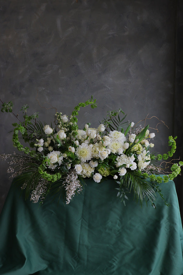 Elegant white and green funeral casket spray with roses, mums, and greenery arranged in a flowing, natural design.