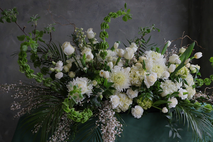 Elegant white and green funeral casket spray with roses, mums, and greenery arranged in a flowing, natural design.