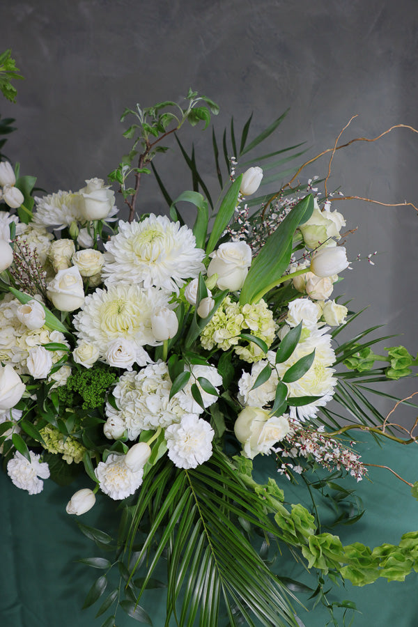 Elegant white and green funeral casket spray with roses, mums, and greenery arranged in a flowing, natural design.