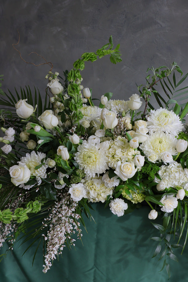 Elegant white and green funeral casket spray with roses, mums, and greenery arranged in a flowing, natural design.