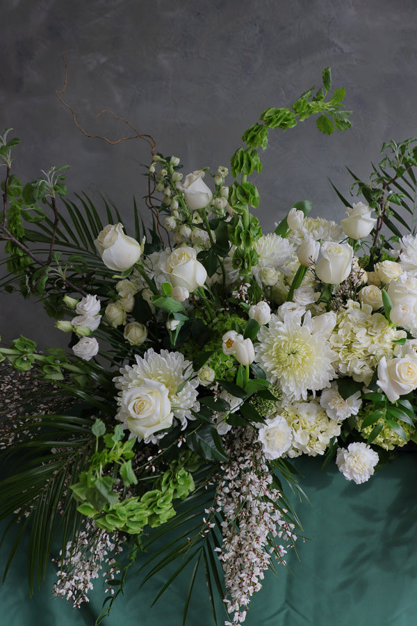 Elegant white and green funeral casket spray with roses, mums, and greenery arranged in a flowing, natural design.