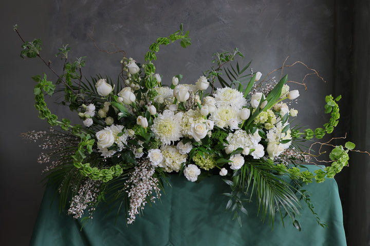 Elegant white and green funeral casket spray with roses, mums, and greenery arranged in a flowing, natural design.