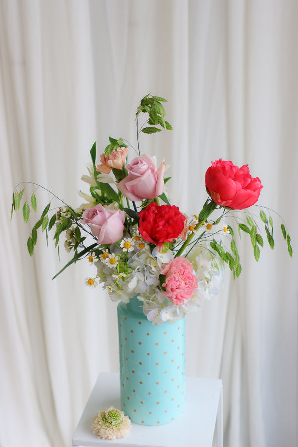 Bright Side flower arrangement with coral peonies, pink roses, blue hydrangeas, and wildflower daisies in a tall blue polka dot vase — cheerful and perfect for any happy occasion.