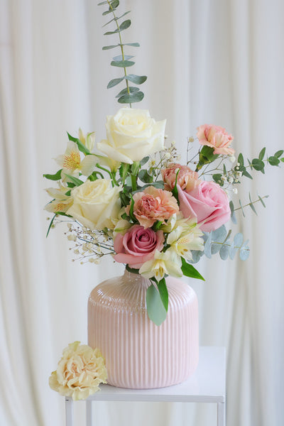 A gentle and delicate floral arrangement featuring blush and white roses with lush white hydrangeas, artfully arranged in an elegant blush ceramic vase. The bouquet exudes warmth, tenderness, and sophistication, perfect for Mother's Day or special occasions.