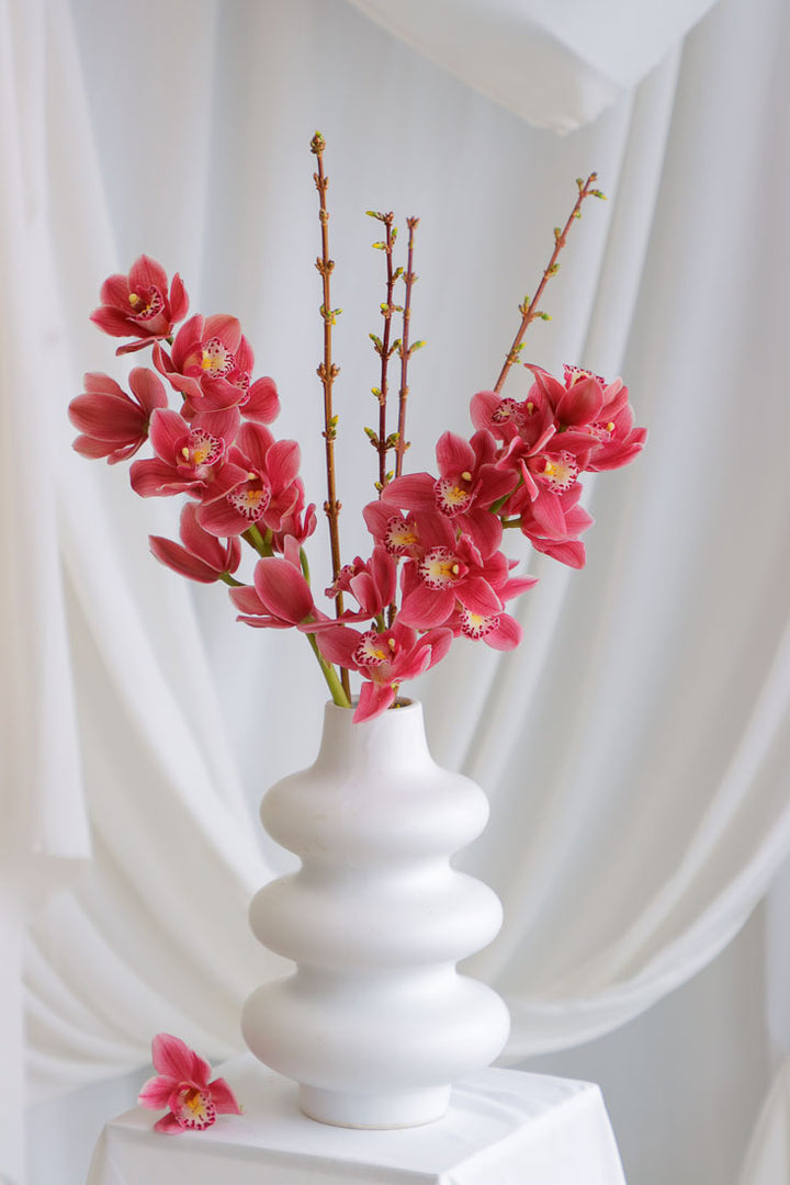 Dark pink mauve Cymbidium orchid arrangement with spring branches in a unique white ceramic vase, styled elegantly.