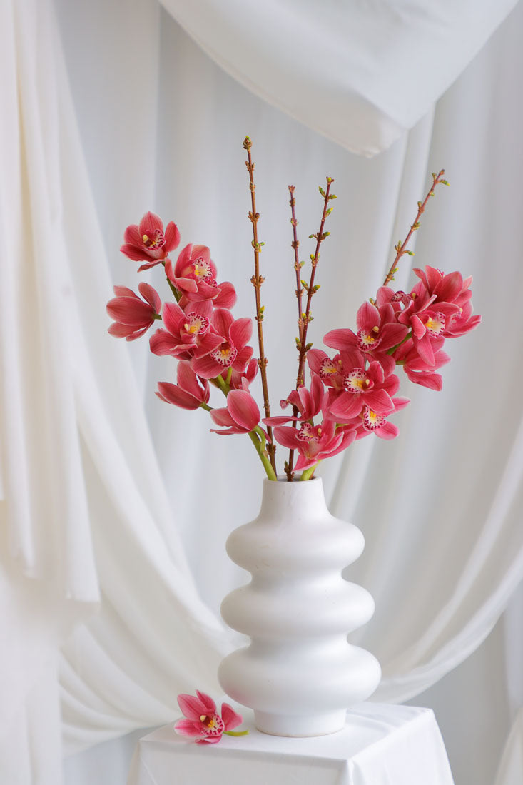 Dark pink mauve Cymbidium orchid arrangement with spring branches in a unique white ceramic vase, styled elegantly.