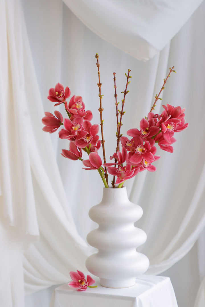 Dark pink mauve Cymbidium orchid arrangement with spring branches in a unique white ceramic vase, styled elegantly.