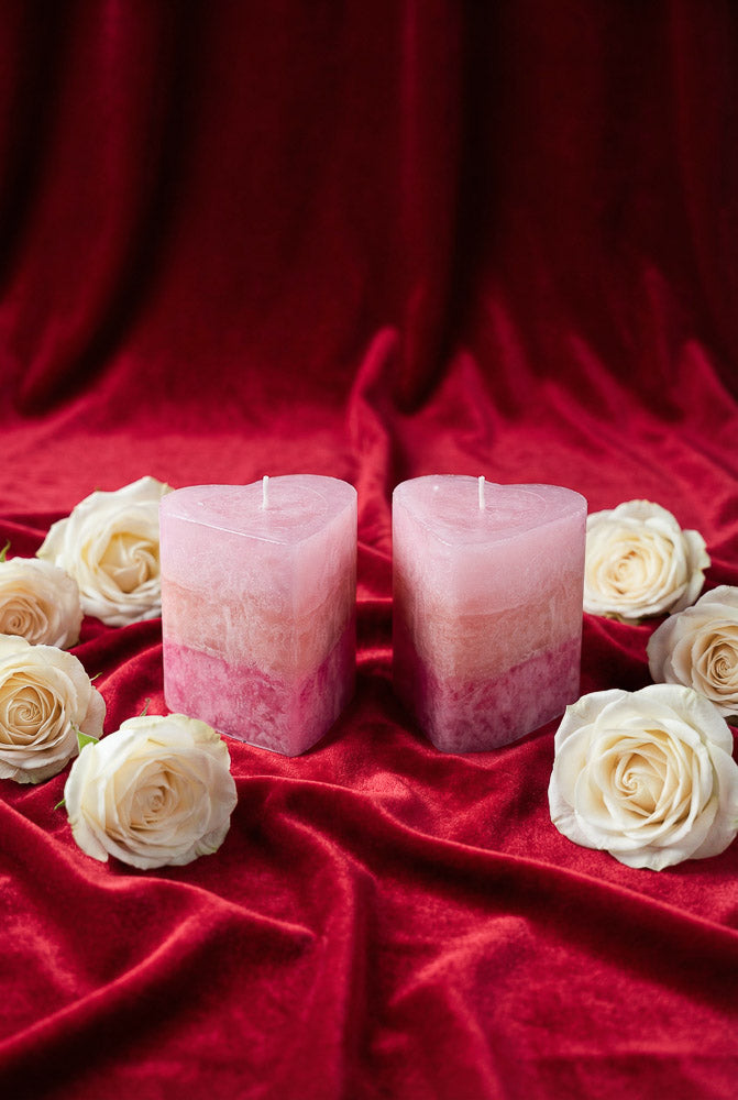 Two blush pink rose-toned pillar candles with a heart-inspired shape, surrounded by white roses on a velvet background for Valentine’s Day