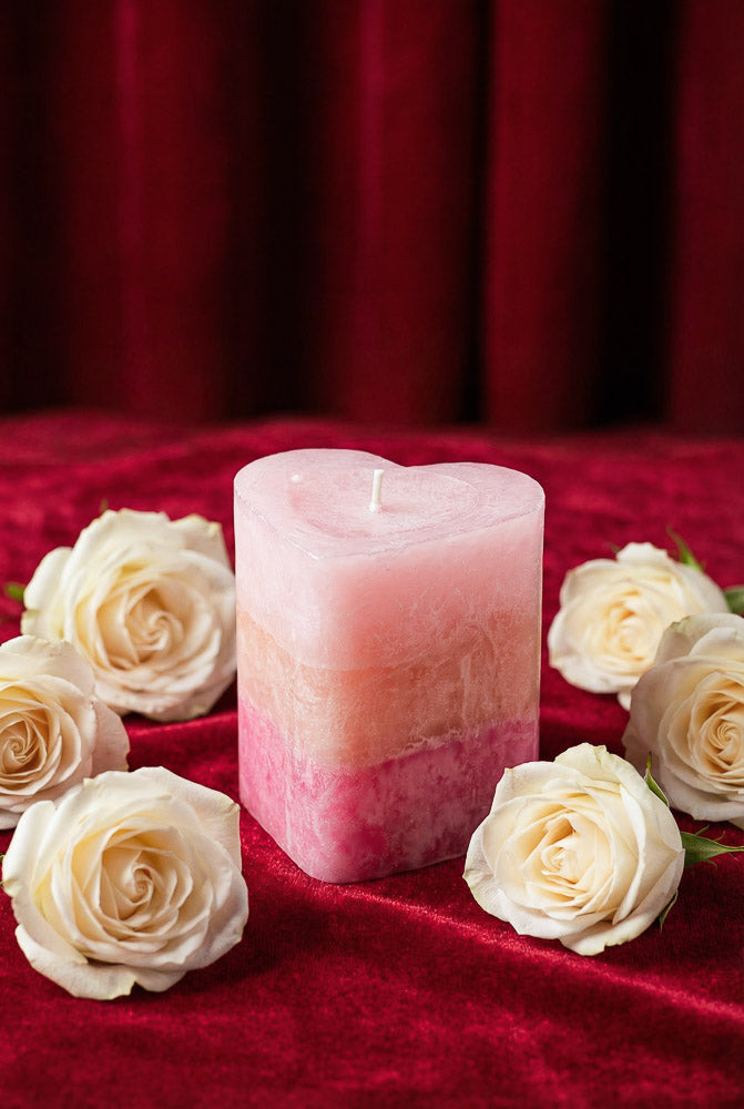 Blush pink rose-toned pillar candle with a heart-inspired shape, surrounded by white roses on a velvet background for Valentine’s Day