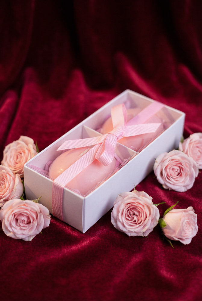 Two pink heart-shaped candles in a gift box with a ribbon, surrounded by blush roses on a velvet background for Valentine’s Day