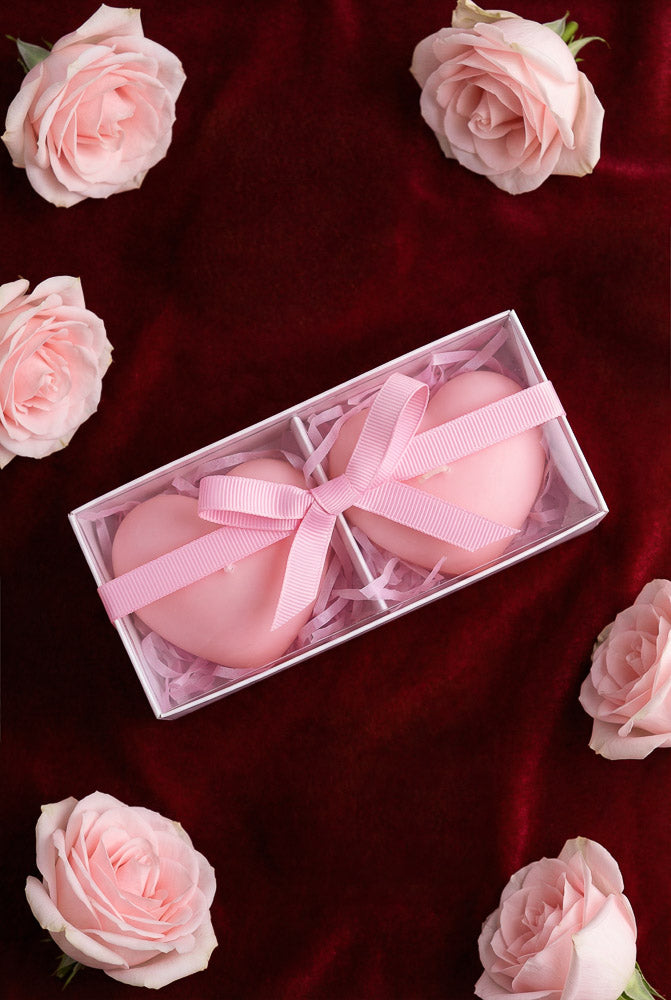 Two pink heart-shaped candles in a gift box with a ribbon, surrounded by blush roses on a velvet background for Valentine’s Day