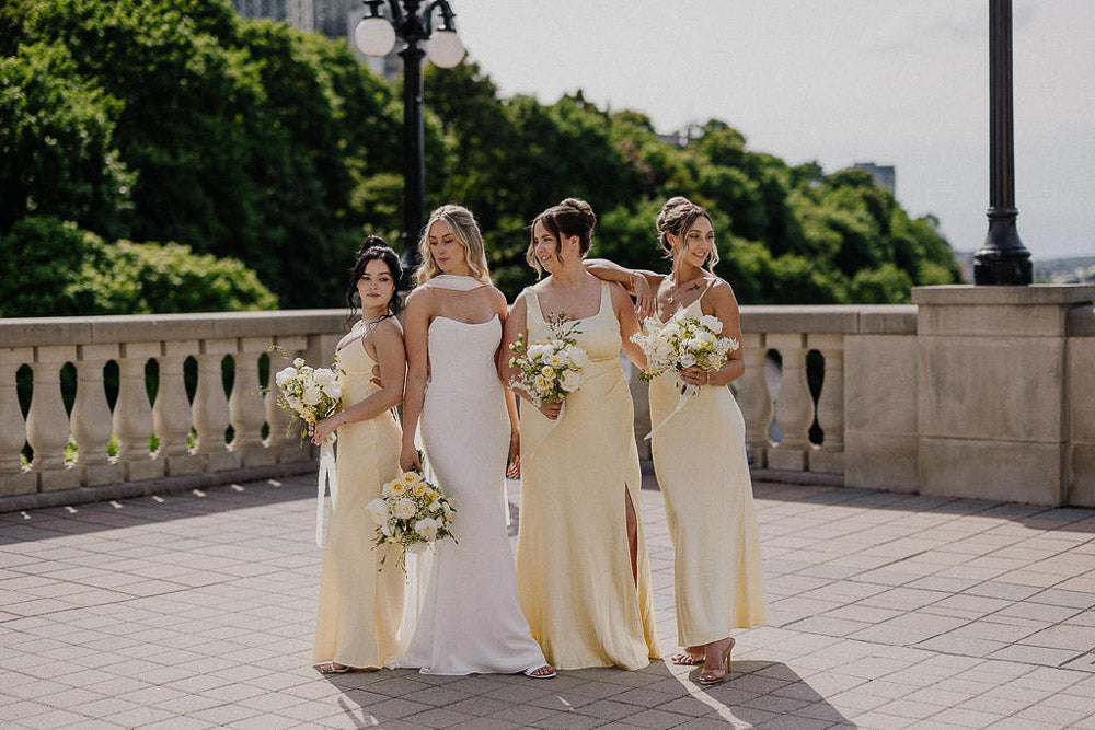 Bridesmaids in light yellow dresses standing together with bridesmaids yellow bouquets on a stone pathway.