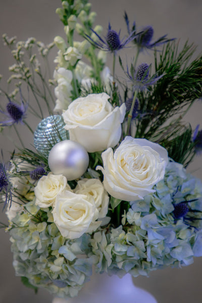 Frosty Grace arrangement with blue and white hydrangeas, roses, snapdragons, thistle, and winter greenery in a modern white ceramic vase by Ottawa florist.