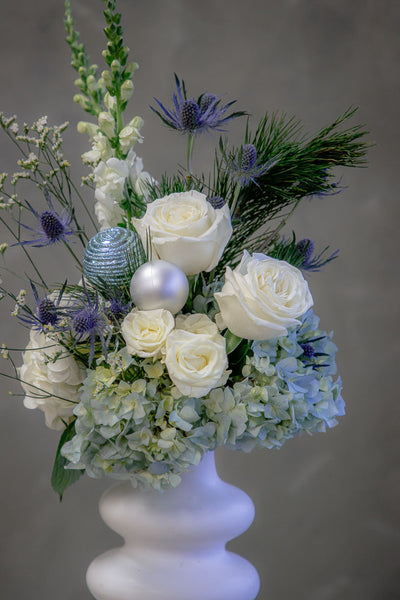 Frosty Grace arrangement with blue and white hydrangeas, roses, snapdragons, thistle, and winter greenery in a modern white ceramic vase by Ottawa florist.