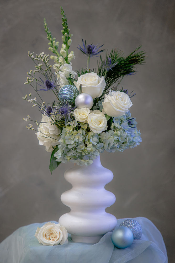 Frosty Grace arrangement with blue and white hydrangeas, roses, snapdragons, thistle, and winter greenery in a modern white ceramic vase by Ottawa florist.