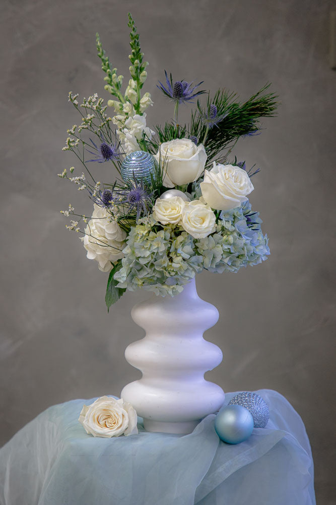 Frosty Grace arrangement with blue and white hydrangeas, roses, snapdragons, thistle, and winter greenery in a modern white ceramic vase by Ottawa florist.