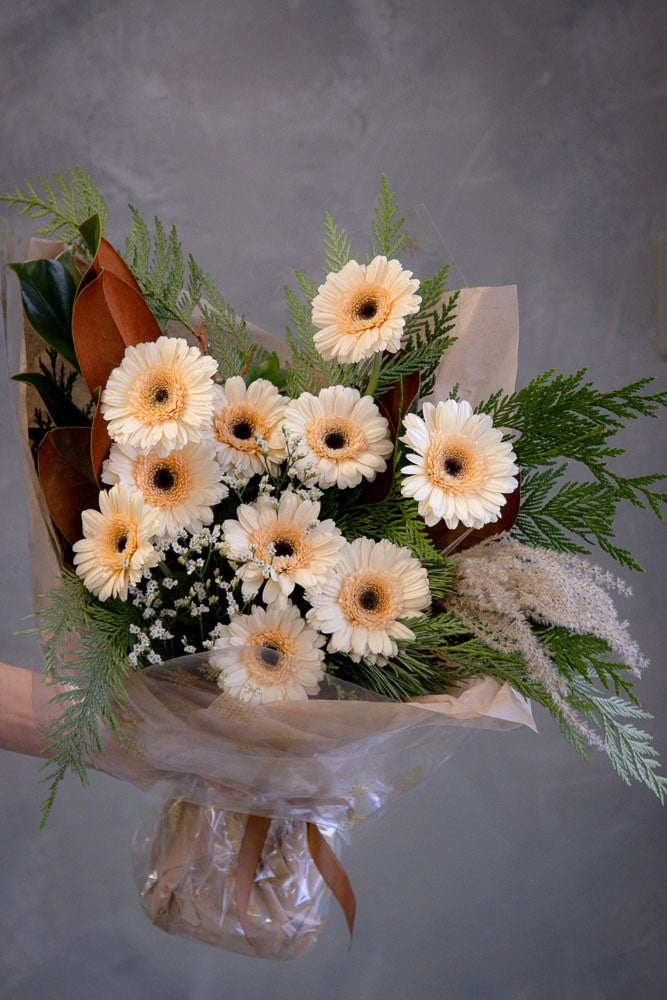 Cozy Winter bouquet with winter foliage, magnolia leaves, dry décor, and peach gerberas wrapped in neutral festive packaging by Ottawa florist.